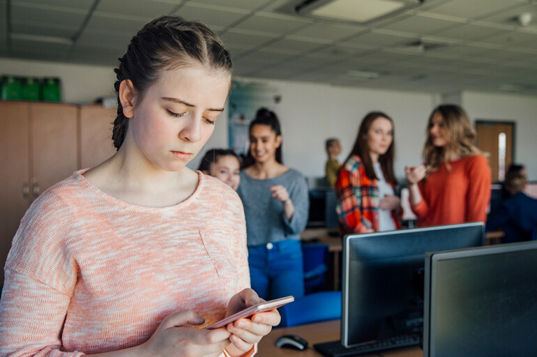 Schoolgirl looking at phone while classmates laugh at her